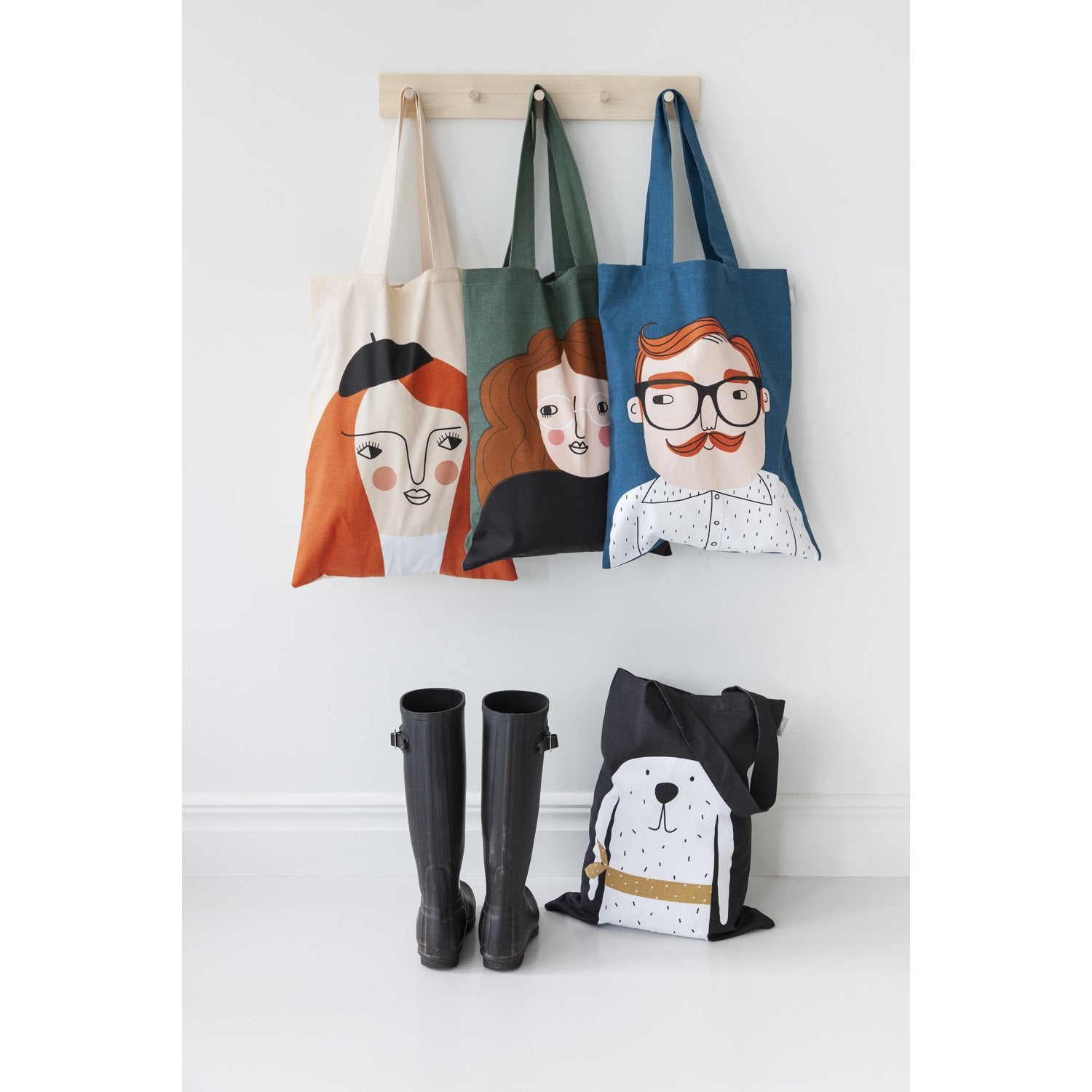 Three tote bags hang on wooden hooks and one tote bag sits on the floor underneath next to a pair of black tall rain boots.