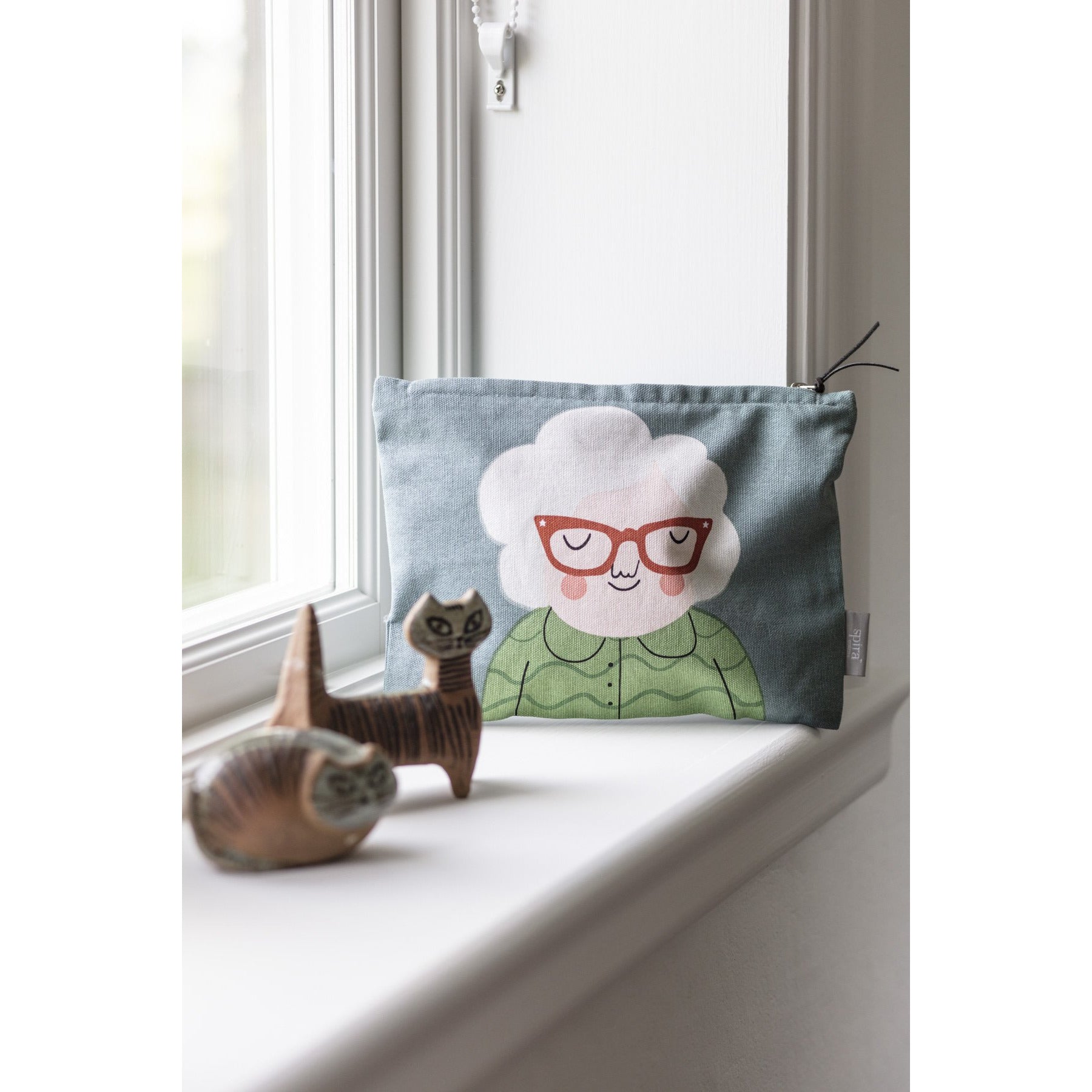 Green gray zipped toiletry bag with women's face with white hair, terra-cotta colored glasses and green shirt. Bag leans against a white windowsill next to two carved wooden cats.