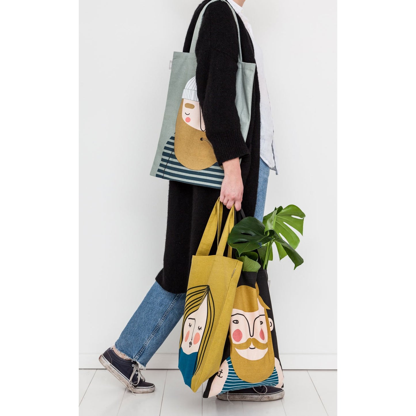 Women in jeans and long black cardigan carrying three tote bags with faces on them.