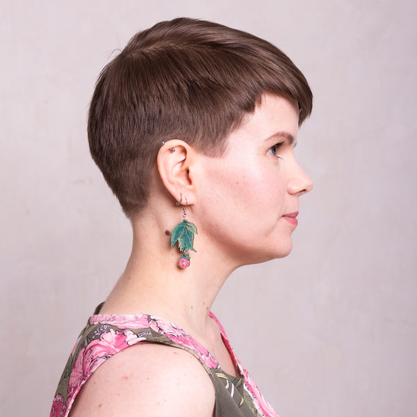 Profile head shot of a woman with brown hair wearing a floral tank top with a leafy green wood earring with small pink peony bud  hanging from ear.
